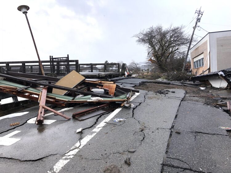 地震や災害によって道路がひび割れ、建物の破片やがれきが散乱している様子。倒れた街灯と傾いた電柱が見える。