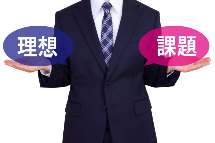 ビジネススーツを着た人物が両手を広げ、左手に『理想』、右手に『課題』と書かれた吹き出しを持っている。