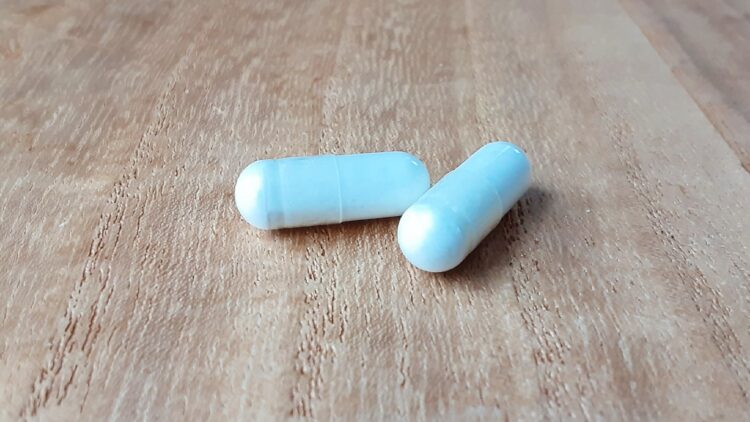 木の机の上に、白いカプセル型のサプリメントが2つ並んでいる。自然な明るさの中で撮影されている。