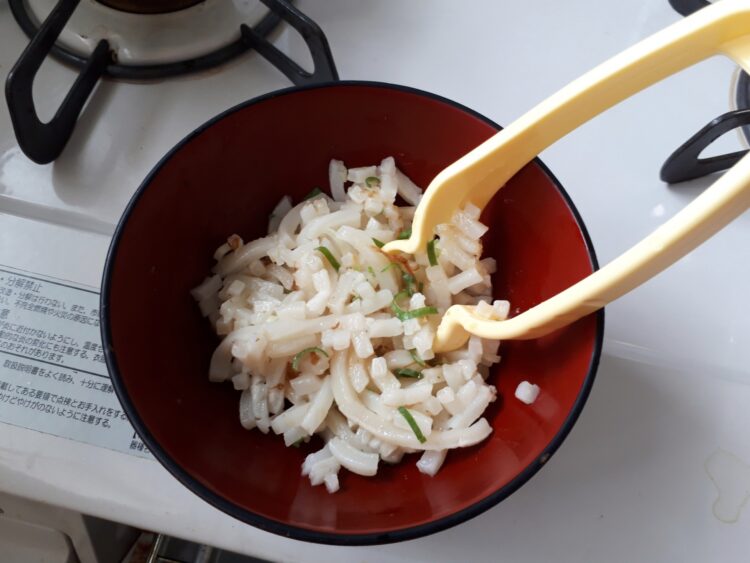 茶色い器に入った炒めたうどん。上に青ねぎがのっていて、黄色いトングでつかまれている。ガスコンロの上に置かれている。