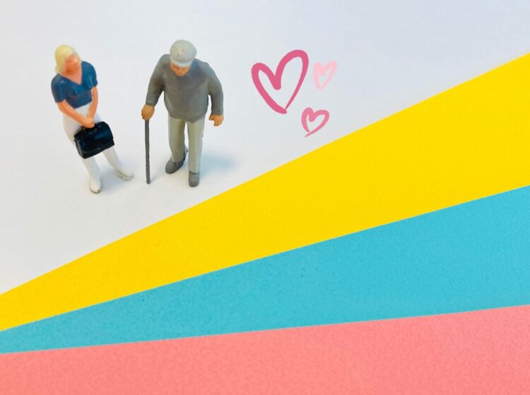 杖をついた高齢の人と若い女性のミニチュアが並んで立っている。背景はピンク・青・黄のカラフルな紙で、そばにハートのイラストが描かれている。