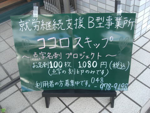 歩道に置かれた緑色の看板に、白い文字で就労支援B型事業所の案内が書かれている。中央に事業所名が大きく書かれ、その下に点字名刺プロジェクトの内容や料金（100枚1080円）などが記されている。下部には利用者募集と電話番号が書かれている。