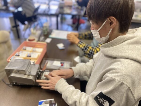 白いパーカーを着た人が、作業机で名刺のようなカードに機械を使って加工している。机の上には加工前のカードが入ったかごが置かれ、奥には他の人も作業をしている様子がある。人物はマスクを着用している