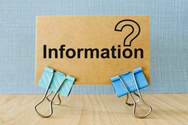 茶色の厚紙に黒い文字で「Information」と書かれ、その右上に大きな疑問符が印刷されている。紙は水色の壁紙を背景に、青と水色の大きなダブルクリップ2つで木目の台の上に立てかけられている。