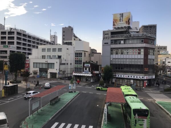 都市のバスロータリーを見下ろした景色。緑色の路線バスが停まっており、交差点を車が行き交う。周囲には中層ビルや商店が並び、上空には青空が広がっている。