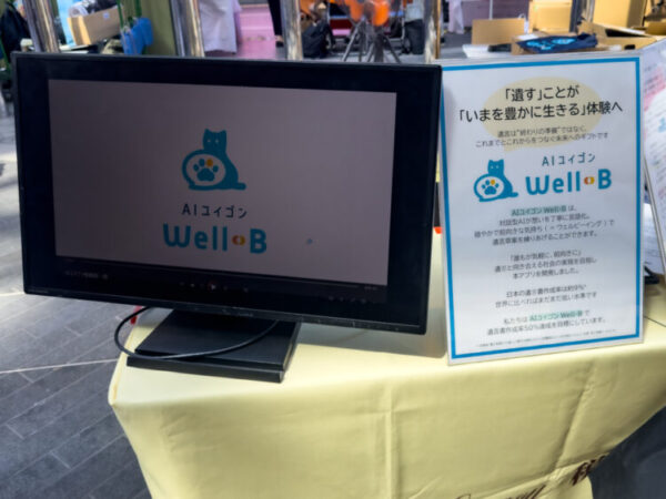 展示ブースの机に置かれたモニターと説明パネル。モニターには猫のイラストと『AIユイゴン Well-B』のロゴが表示されている。隣のパネルには、『遺すことがいまを豊かに生きる体験へ』というキャッチコピーとサービスの説明文が書かれている