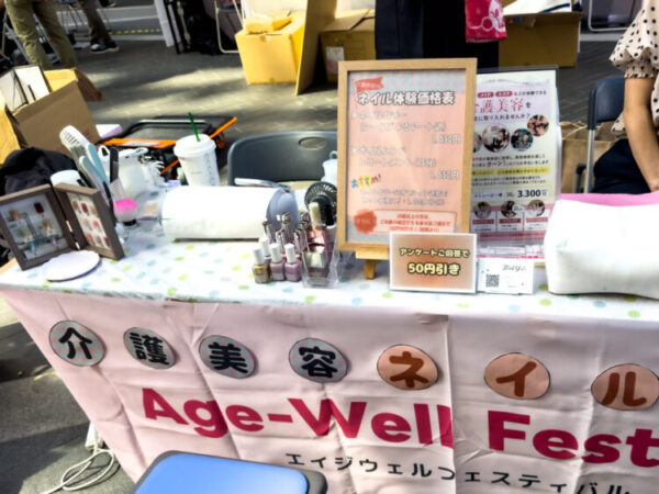 Age-Well Festival の会場に出展された『介護美容ネイル』の体験ブース。机の上にはネイルポリッシュや道具が並び、価格表にはネイルカラーやハンドトリートメントの体験料金が示されている。机の前には『介護美容ネイル』と書かれた布が掛けられている。