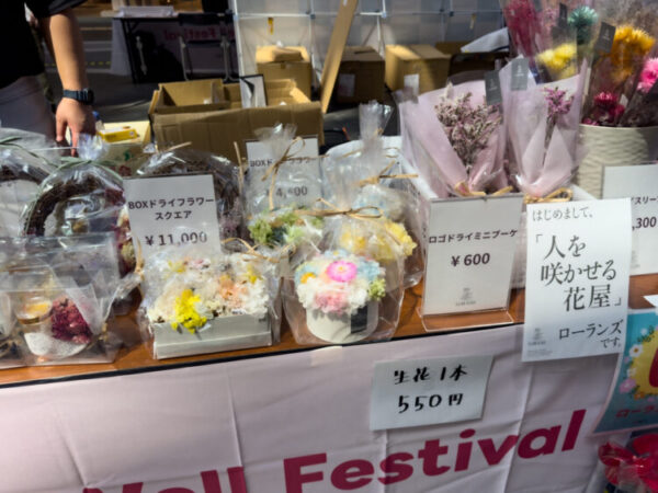Age-Well Festival 2025 の会場に出展された花屋ローランズのブース。ドライフラワーのリースやミニブーケ、カラフルな生花が並び、値札には600円から11,000円までの価格表示がある。『人を咲かせる花屋』という紹介文が掲示されている。
