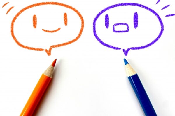 白い紙に、オレンジ色と紫色のクレヨンで描かれた吹き出し型の顔が二つ。左は笑顔、右は口を丸く開けた驚いた表情。下にはそれぞれ同じ色の色鉛筆が置かれている。