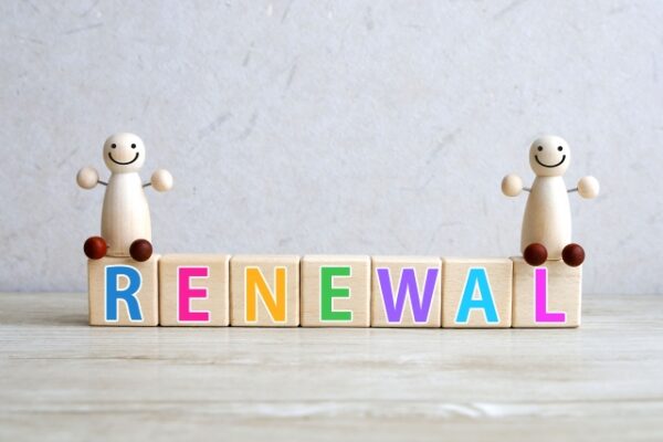 カラフルな文字で「RENEWAL」と書かれた木のブロックが横に並び、両端に笑顔の木製人形が座っている。背景は淡いグレーの壁で、テーブルの上に置かれている。