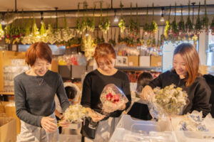 花屋の作業場で3人の女性が笑顔でドライフラワーの花束を作っている。 それぞれの手元には白やピンクの花があり、丁寧にラッピングをしている様子。 背景には天井から吊るされた多数のドライフラワーが彩り豊かに並び、あたたかい照明が作業場を柔らかく照らしている。 楽しそうに協力しながら花を束ねる、穏やかな雰囲気の職場風景。