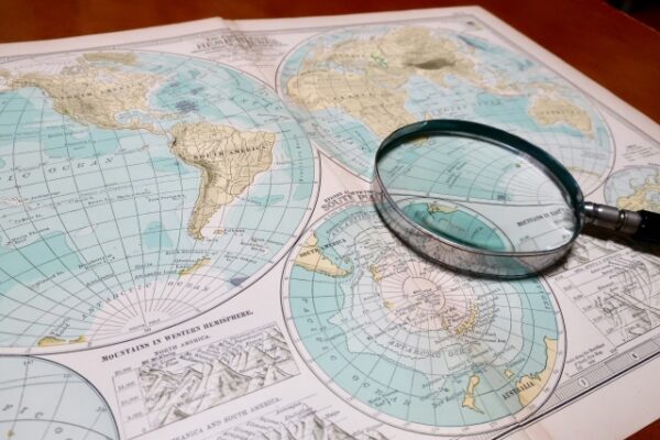 世界地図が描かれたページの上に、銀色の虫眼鏡が置かれている。地図は青とベージュを基調とし、地理や探検、調査をイメージさせる構図。