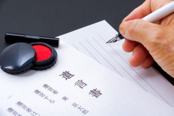 遺言書の用紙に万年筆で文字を書こうとしている手。横には赤い印鑑の朱肉とキャップが置かれており、正式な書類作成の場面を表している。