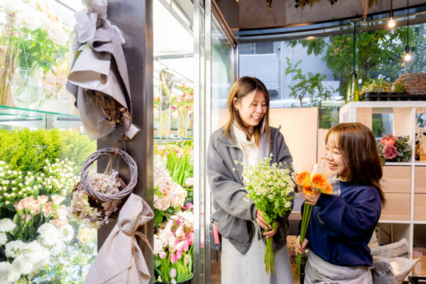 店内の花屋で、2人の女性が笑顔で会話をしながら花束を手にしている。 1人は白い小花のブーケを、もう1人はオレンジの花を持ち、楽しそうに並んで立っている。 周囲には色とりどりの花々が並び、ガラス越しに自然光が差し込む明るい空間。 生花の香りと温かな雰囲気が伝わる、和やかな接客の様子。