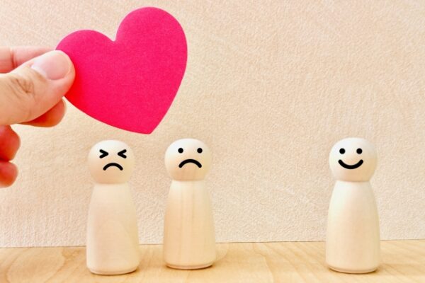 3体の木製人形が並び、左から順に怒った顔、悲しい顔、笑顔の表情をしている。左上には手で持たれた赤いハートの紙があり、感情や思いやりを表すイメージ。