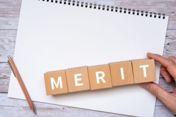 木製のブロックを横に並べて「MERIT」と白い文字で書かれている。背景にはノートと鉛筆が置かれ、手がブロックを押さえている。利点やメリットを表すイメージ。