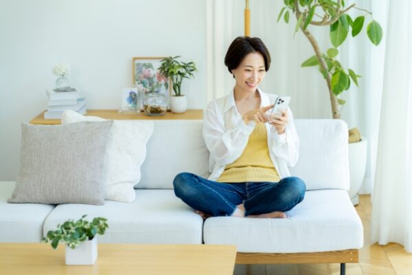 明るいリビングで、ソファに座った女性がスマートフォンを見ながら笑顔で操作している。周囲には観葉植物や写真立てが置かれ、穏やかな日常の雰囲気が伝わる。