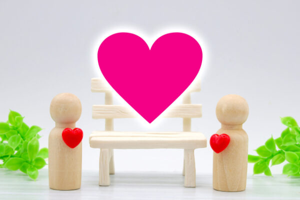 木製の人形が2体、向かい合って立っており、それぞれ胸に赤いハートの飾りをつけている。中央のベンチの上には大きなピンク色のハートが浮かび、愛情や絆を表現している。