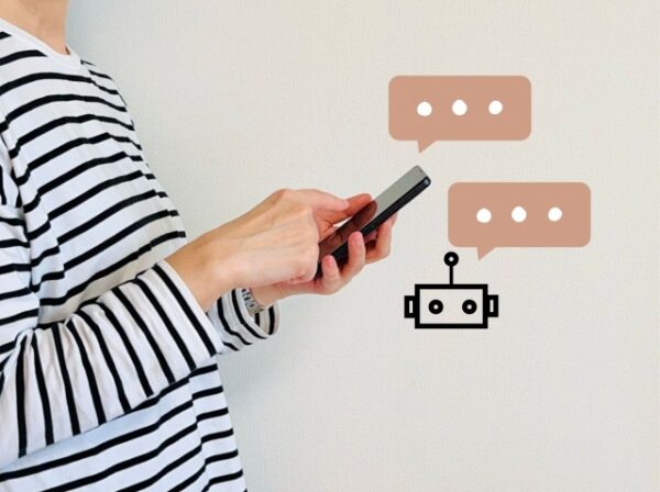ボーダーの服を着た人がスマートフォンを操作している。壁の前に立ち、画面の横には吹き出しと小さなロボットのイラストが描かれており、チャットボットとの会話を表している。