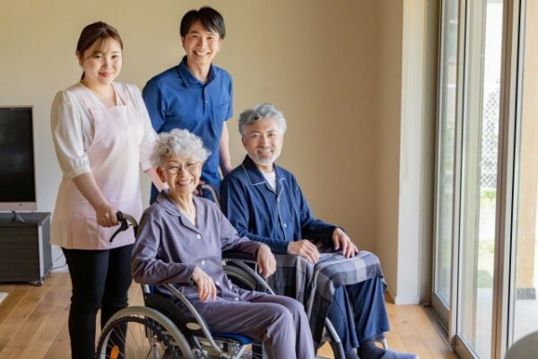 明るい室内で、車いすに座る高齢の女性と、椅子に座る高齢の男性が笑顔で並んでいる。後ろには介護士の男女が立ち、全員が和やかな表情でカメラを見ている。家庭的な介護の様子を表す写真。