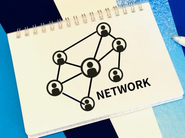 白いノートに、複数の人のアイコンが線でつながったネットワーク図が描かれている。右下には「NETWORK」という文字。背景は青と白の紙が重なり、デジタルなつながりや協力をイメージした構成。