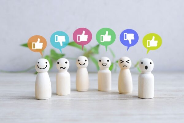 7体の木製人形が横一列に並び、それぞれ異なる表情をしている。頭の上には親指の上げ下げアイコンが描かれたカラフルな吹き出しがあり、人の感情や意見の違いを表している。