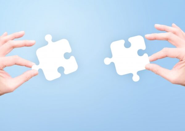 青い背景の中で、左右から伸びた2本の手が、それぞれ白いジグソーパズルのピースを持っている。2つのピースは向かい合っており、つながりや協力を表現している。