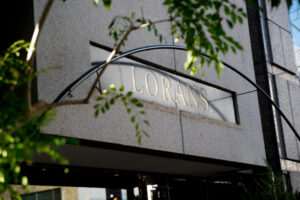 建物の外壁に「LORANS.」と刻まれた看板が掲げられている。 周囲には緑の葉がやわらかくかかり、自然と調和した落ち着いた雰囲気。 日差しを受けて金色の文字が輝き、洗練された外観が印象的な店舗入口の一部。