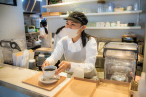カフェのカウンターで、マスクをつけた女性スタッフが笑顔でコーヒーを差し出している。 スタッフは白いシャツとエプロン、黒いキャップを着用し、温かみのある雰囲気の店内で接客中。 背後にはキッチンと棚に整然と並んだカップやグラスがあり、清潔感のある明るい空間が広がっている。 心のこもったサービスと穏やかな笑顔が印象的なワンシーン。
