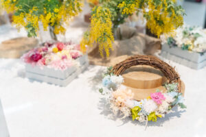 白いテーブルの上に、色とりどりのドライフラワーやリースが並べられている。 手前には淡いピンクやブルーの花をあしらったリースが置かれ、奥には黄色いミモザの花が華やかに飾られている。 木製の台やナチュラルな布が添えられ、やさしく明るい雰囲気のディスプレイ。 自然の温もりと季節感が感じられる、花のアトリエの一角。