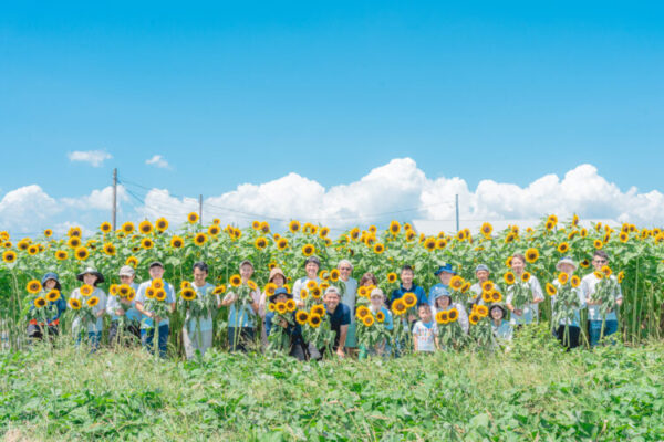 一面に咲くひまわり畑の前で、20人ほどの人々が笑顔で並んで記念撮影をしている。 真っ青な空と白い雲が広がる夏の日差しのもと、それぞれが手にひまわりを持ち、明るく爽やかな雰囲気に包まれている。 自然と人の温かさが感じられる集合写真。