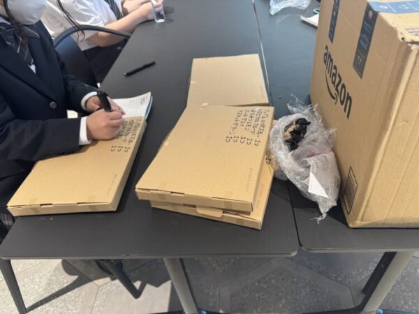 ChatGPT:altテキスト案 制服姿の学生がテーブルに座り、段ボール製の平たい箱にマジックペンでメモを書いている。 隣にはAmazonの段ボール箱や梱包材が置かれており、ワークショップのような作業風景が広がっている。