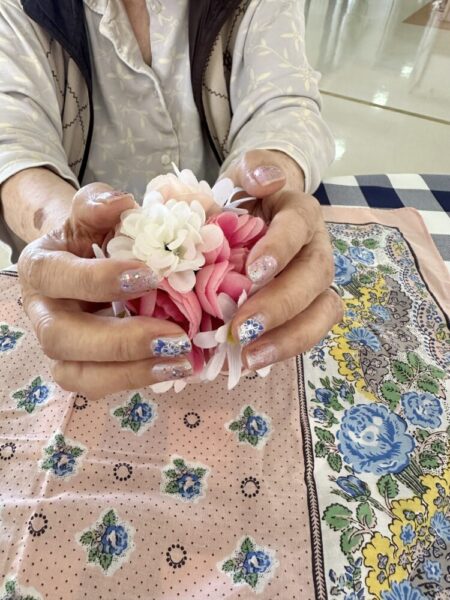 高齢の女性が、ピンクと白の布製の花を両手でやさしく持っている。爪にはラメや花柄のネイルアートが施され、華やかな印象。テーブルには花柄のハンカチが敷かれている。