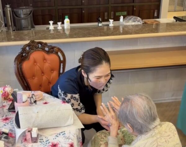 室内で、若い女性が高齢者の手を優しく握りながらネイルや手のケアをしている。テーブルには化粧品やネイル用品が並び、介護現場での美容サービスの一場面を表している。