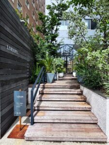 木製の階段を上ると、緑に囲まれたガラス張りの建物が見える。 左側の黒い木製フェンスには「LORANS.」の文字があり、入り口手前には営業時間を示す小さな看板が立てられている。 植物に包まれた小道のようなアプローチが印象的で、自然と調和した落ち着いた雰囲気の外観。
