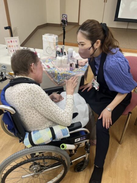 車いすに座る高齢の女性が、テーブル越しに美容スタッフと向き合っている。スタッフはマスクをつけ、女性の手を取りながら優しく会話をしている。テーブルの上にはネイル用品やケア道具が並び、温かい雰囲気で美容サービスが行われている。