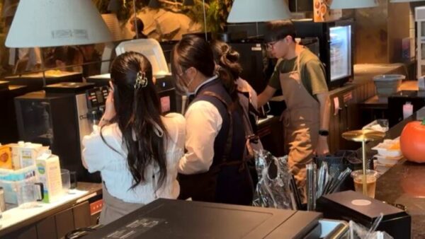 カフェのカウンター内で、3人のスタッフが並んで作業している様子。 一人はエスプレッソマシンの前で飲み物を準備し、もう一人は指導するようにそばで見守っている。 男性スタッフも後方でドリンク作りに取り組んでおり、店内は温かい照明に包まれている。