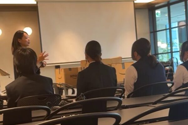 教室のような会議室で、講師が前方で話しながら手を動かして説明している。 受講者の数名が椅子に座って講師の話を聞いており、正面にはホワイトスクリーンと段ボール箱が見える。 明るい室内で、学びの雰囲気が感じられる場面。