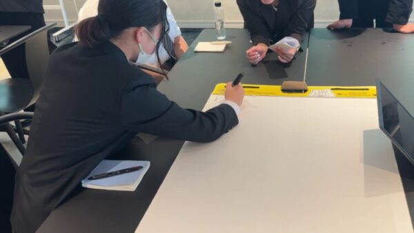 スーツ姿の学生たちがテーブルを囲み、模造紙にアイデアを書き込んでいる。 中央の学生がマーカーで文字を書き、周囲の学生はノートを持ちながら意見を出している。 グループワークのような協働作業の様子がうかがえる。