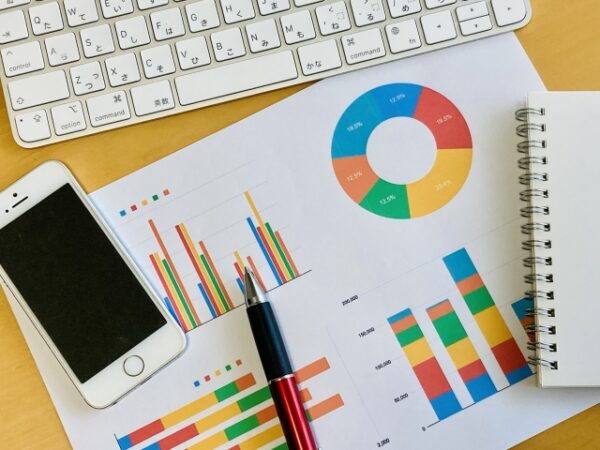 木目の机の上に、グラフが印刷された数枚の紙が置かれている。 円グラフ、棒グラフ、積み上げグラフなどがあり、赤・青・黄・緑などの色で示されている。 その上に黒と赤のボールペンが置かれ、左側には白いスマートフォン、上部には白いキーボード、右側には小さなノートがある。 ビジネスやデータ分析、報告書づくりの場面をイメージさせる構図。全体的に明るい光が差し込んでおり、清潔で整理された印象の写真。