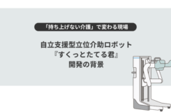 淡いグレーの背景の中央に、黒い文字で大きく「自立支援型立位介助ロボット『すくっとたてる君』開発の背景」と書かれている。 その上には黒い帯に白文字で「持ち上げない介護」で変わる現場、という見出しがある。 右側には白と水色を基調とした介助ロボットのイラストが描かれ、立ち上がりをサポートする形状が示されている。 全体として、介護の負担軽減や自立支援をテーマにしたスライドのタイトル画像。