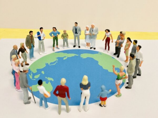 地球の形をした大きな円の上に、さまざまな人々の小さな人形がぐるりと輪になって立っている。 人形は男女・年齢・人種などがさまざまで、スーツ姿の人、子どもを抱く人、杖をつく高齢の人などがいる。 地球の模様は青と緑で描かれ、アジアやオセアニアのあたりが見えている。 世界中の人がつながり、共に生きることをイメージした温かい印象の写真。