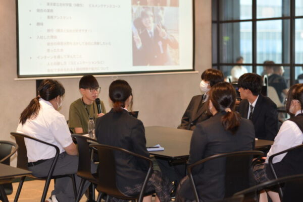 会議室の一角で、数人の高校生が円卓を囲み、スーツや制服姿で話を聞いている。 中央では、眼鏡をかけた人物がマイクを持って話しており、落ち着いた雰囲気で質疑応答をしているようす。 背後のスクリーンには発表スライドが映し出され、仕事内容や入社理由、インターンで学んだことなどが書かれている。 窓の外にはビルの風景が見え、現場のリアルな就業体験を共有するような場面を写している。