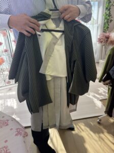 ストライプ柄の青いシャツを着た男性が、和装の衣装をハンガーにかけて両手で持っている。 衣装は緑色の縦じま模様の羽織と、白い着物、灰色の帯で構成されている。 背景には柔らかな光が差し込み、観葉植物や淡いピンク色の花が見える。 和装を丁寧に紹介しているような、落ち着いた雰囲気の室内で撮影された写真。