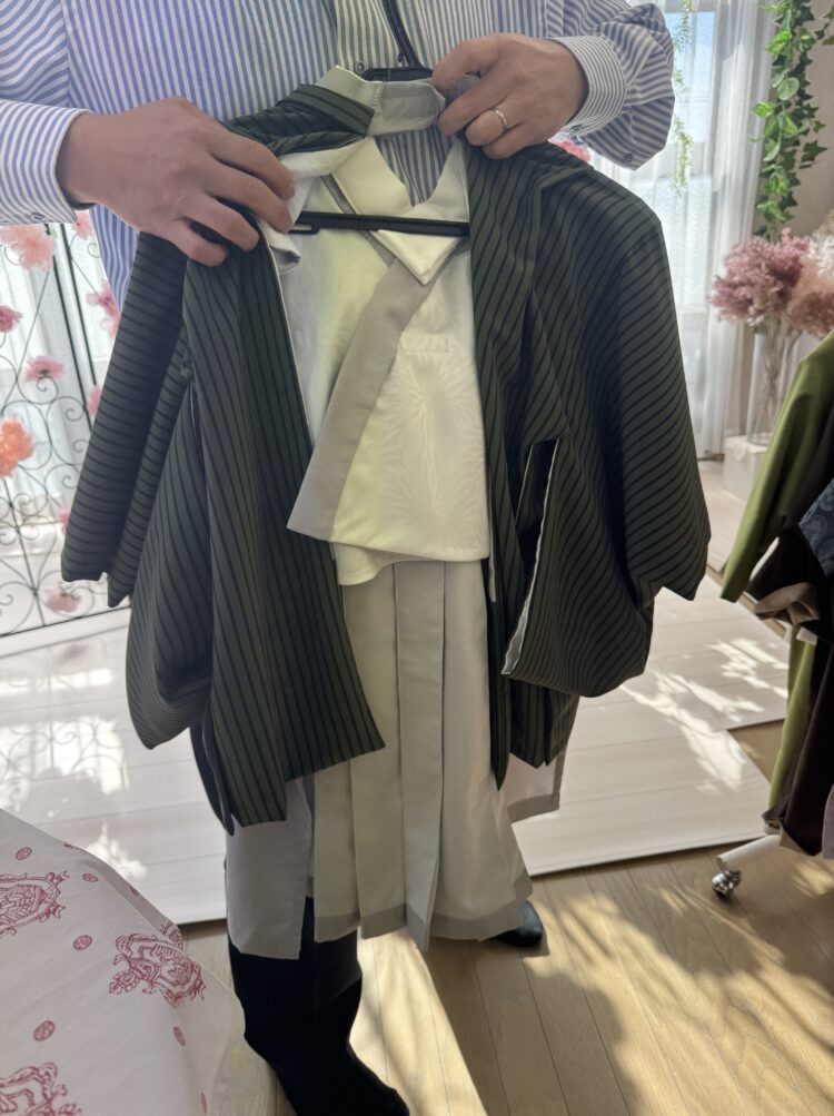 ストライプ柄の青いシャツを着た男性が、和装の衣装をハンガーにかけて両手で持っている。 衣装は緑色の縦じま模様の羽織と、白い着物、灰色の帯で構成されている。 背景には柔らかな光が差し込み、観葉植物や淡いピンク色の花が見える。 和装を丁寧に紹介しているような、落ち着いた雰囲気の室内で撮影された写真。