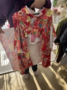 紫色の服を着た人が、赤い着物をハンガーにかけて両手で持っている。 着物には花や車輪などの華やかな和柄が描かれており、黄色・桃色・水色など多彩な色が使われている。 白い襟とピンクの帯が見え、全体的に明るく鮮やかな印象。 背景には柔らかな日差しが差し込み、観葉植物やドライフラワーが並ぶ落ち着いた室内で撮影された写真。