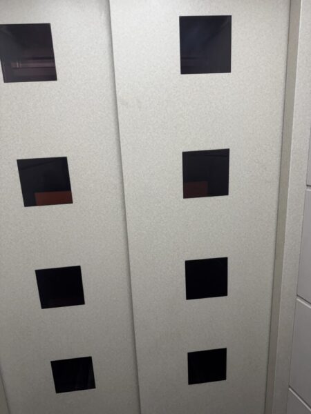 明るい灰色の金属製のドアが映っている。 ドアは中央で左右に分かれており、両側に縦に3つずつ、計6つの黒い四角い窓が並んでいる。 周囲は白っぽい壁で囲まれ、全体的にシンプルで清潔な印象。 建物のエレベーターの扉を正面から撮影した写真。