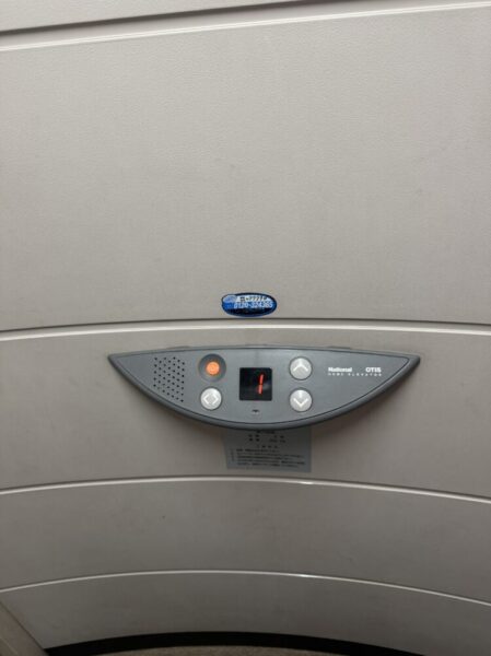 エレベーター内部の操作パネルが壁に取り付けられている。 壁は明るいベージュ色で、中央のパネルは灰色の半円形をしており、左右に開くボタンと下向き・上向きの矢印ボタンが配置されている。 中央の小さな表示画面には赤い数字の「1」が点灯しており、現在の階数を示している。 全体的にシンプルで清潔なデザインの家庭用エレベーター内の様子。