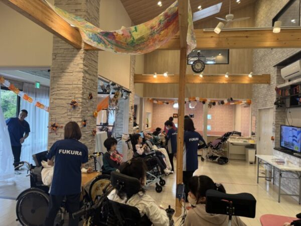 明るい室内で、数人のスタッフと車いすを利用する利用者たちが過ごしている。 スタッフは青いTシャツを着ており、背中に「FUKURA」と書かれている。 壁や天井にはカボチャやオレンジ色の飾りがあり、ハロウィンの装飾が施されている。 天井が高く、木の梁や白い壁があり、開放的で温かい雰囲気の空間。 スタッフが利用者のそばに寄り添いながら、穏やかに交流している様子が伝わる写真。