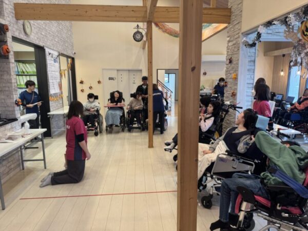 広い室内で、複数の車いすの利用者が並び、スタッフと一緒に活動している。 中央にはピンク色の制服を着た職員が膝をついて話しかけており、周囲には青い制服のスタッフもいる。 壁や天井にはハロウィンの飾りつけがされ、カボチャやオレンジのガーランドが見える。 利用者たちは笑顔で職員と交流しており、にぎやかで温かい雰囲気のある場面。